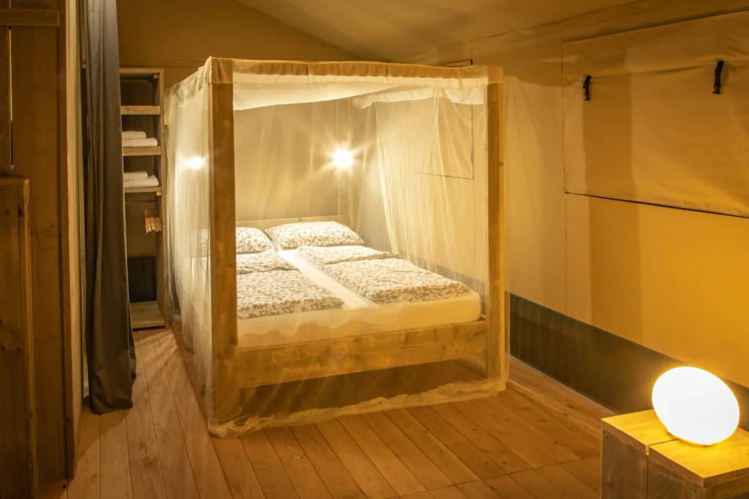 Chambre du Wood Lodge pouvant accueillir de 2 à 6 personnes, avec un lit à baldaquin sous une moustiquaire, une armoire ouverte contenant des serviettes et une lampe sur la table de chevet