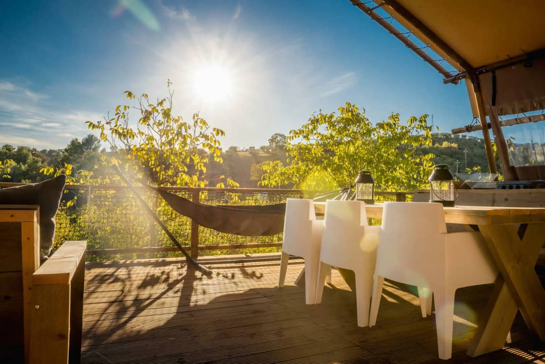 Table à manger en bois à pieds croisés et chaises blanches sur la terrasse du Desert Lodge XL, avec le soleil au-dessus des arbres