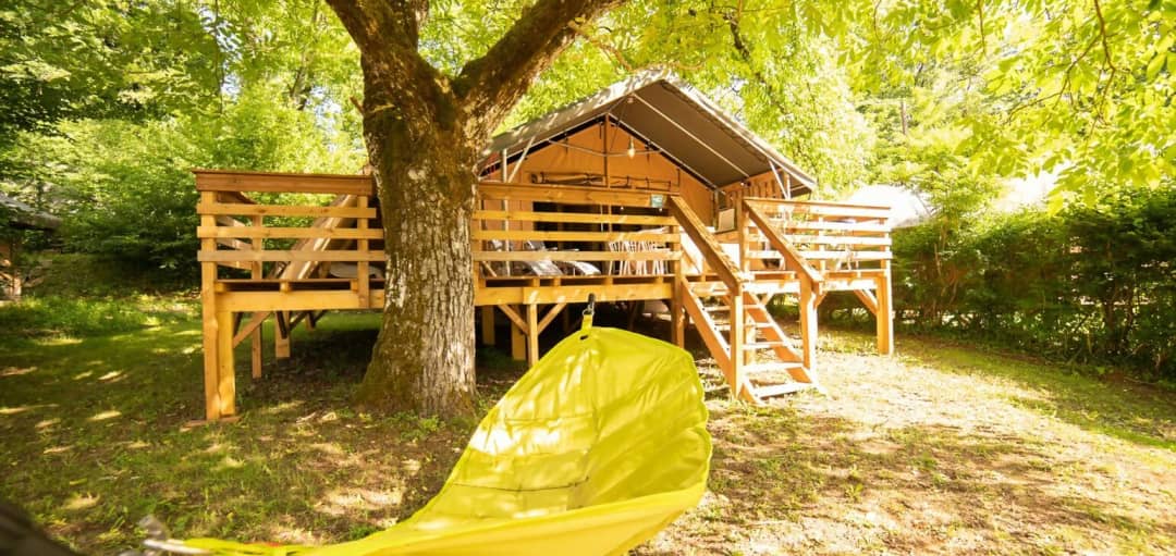 Lodge de safari sur pilotis sous un grand arbre, avec un escalier en bois menant à la terrasse et un hamac jaune au premier plan