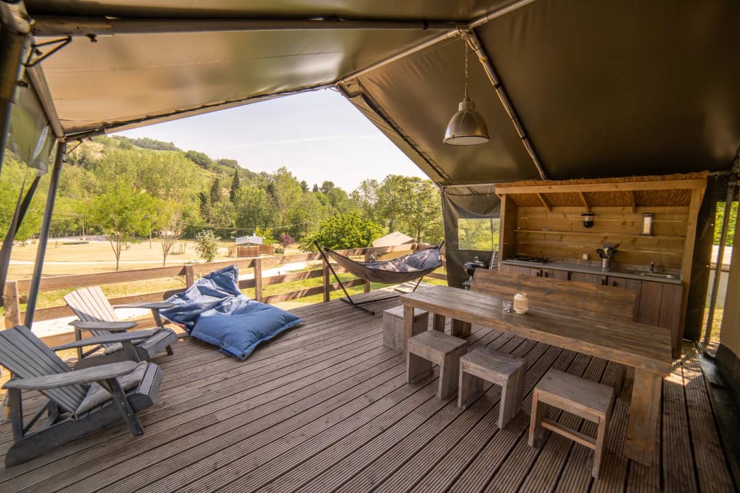 Véranda du Safari Lodge pouvant accueillir de 2 à 6 personnes, avec des chaises de jardin, des poufs bleus, un hamac et une longue table avec des tabourets, surplombant une pelouse