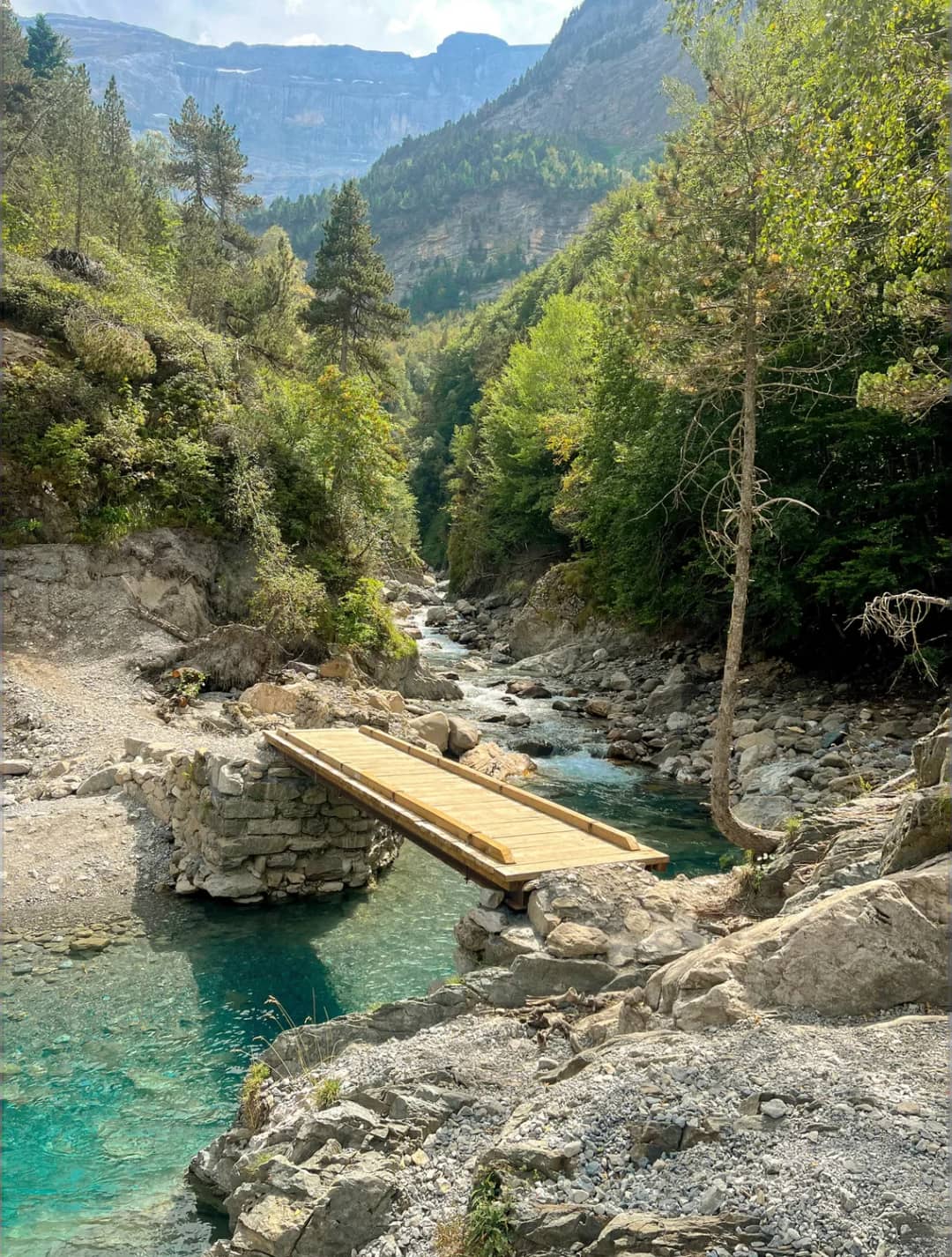 Rivière de montagne d'un bleu limpide avec plateforme en bois dans une vallée boisée entourée de rochers et de conifères, montagnes en arrière-plan.